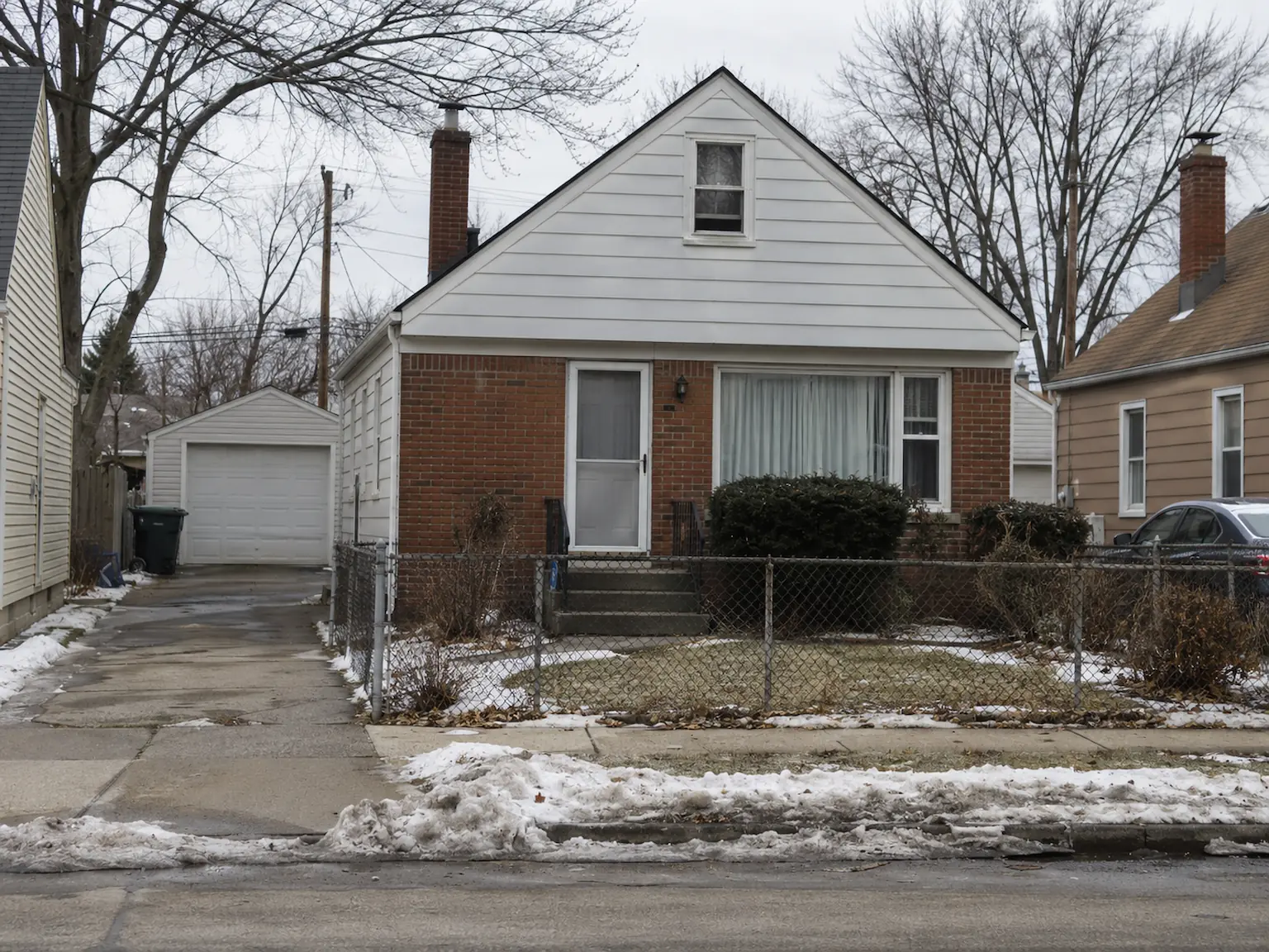 Post-war brick Cape Cod bungalow with a detached garage and chain-link fence in a Cleveland neighborhood in winter