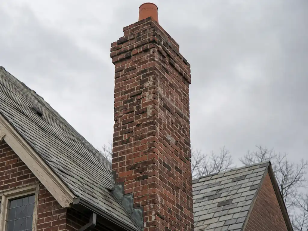 Tall brick chimney with deteriorating mortar joints rising through a slate roof on a Heights Tudor