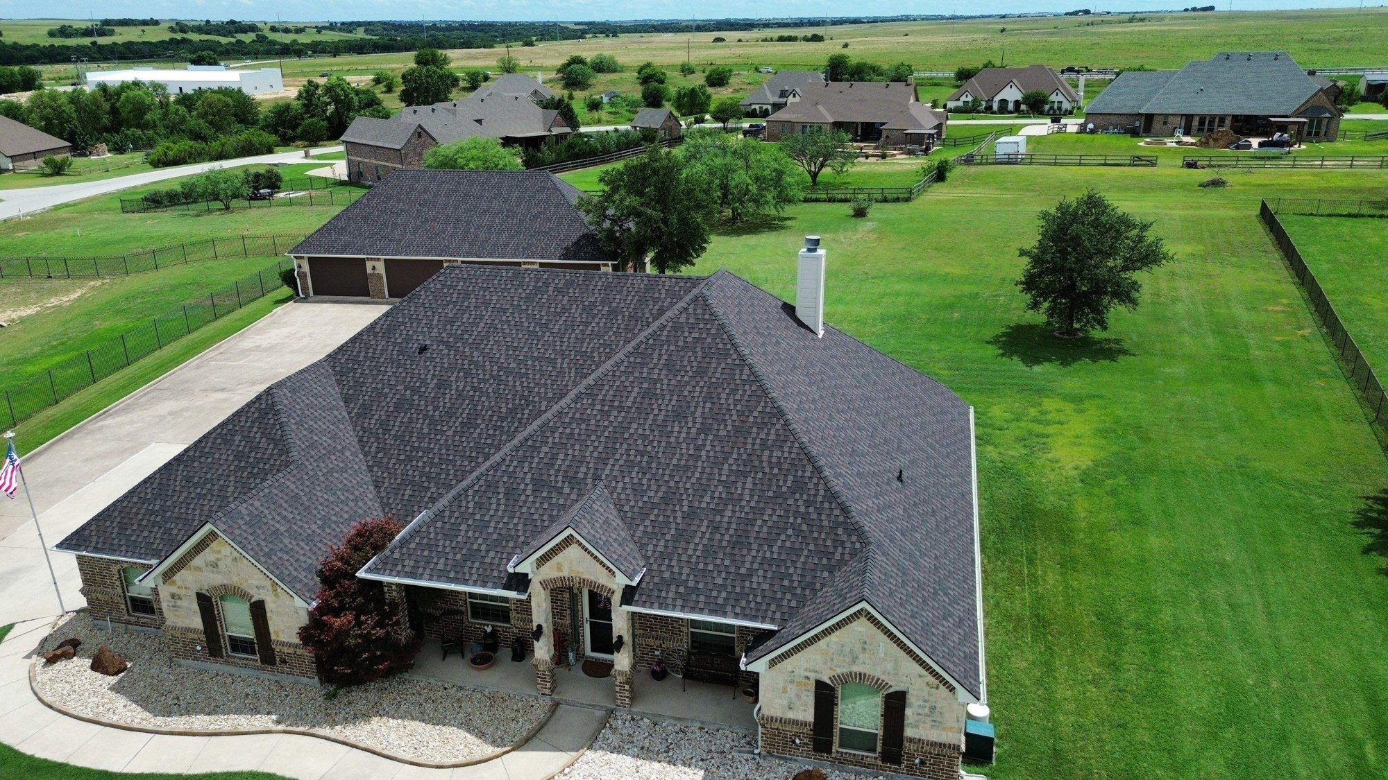 Drone view of a finished dark architectural shingle roof on a brick and stone home with a flag out front in Farmers Branch, TX