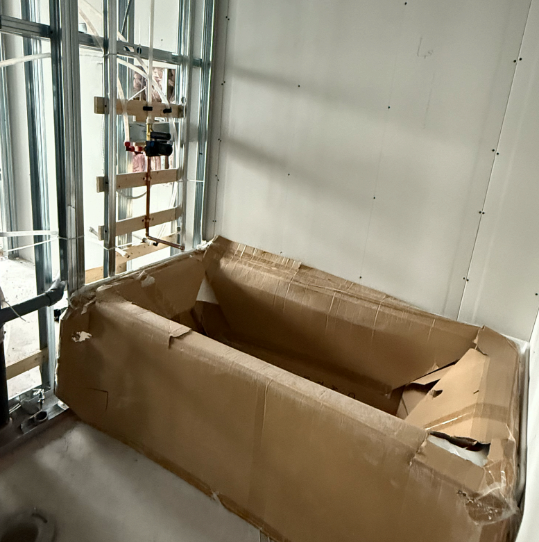 Bathtub set in place and wrapped in cardboard protection, with copper pipework and a mixing valve roughed in on the wall behind