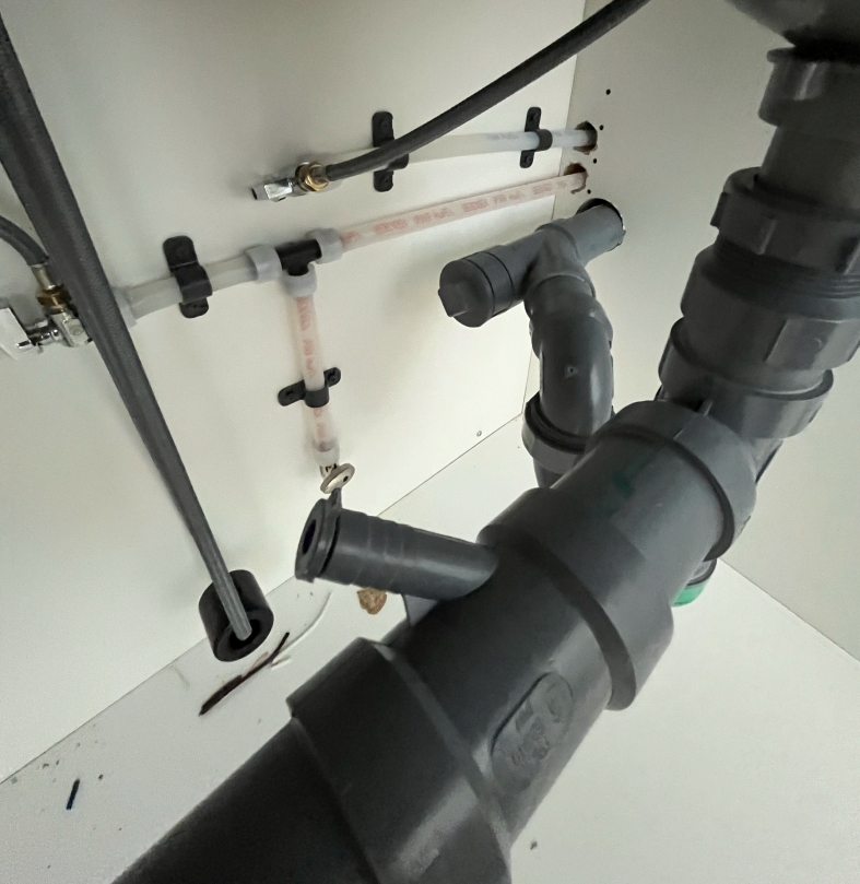 Double-bowl stainless kitchen sink from below, showing continuous waste pipework and a food waste disposal