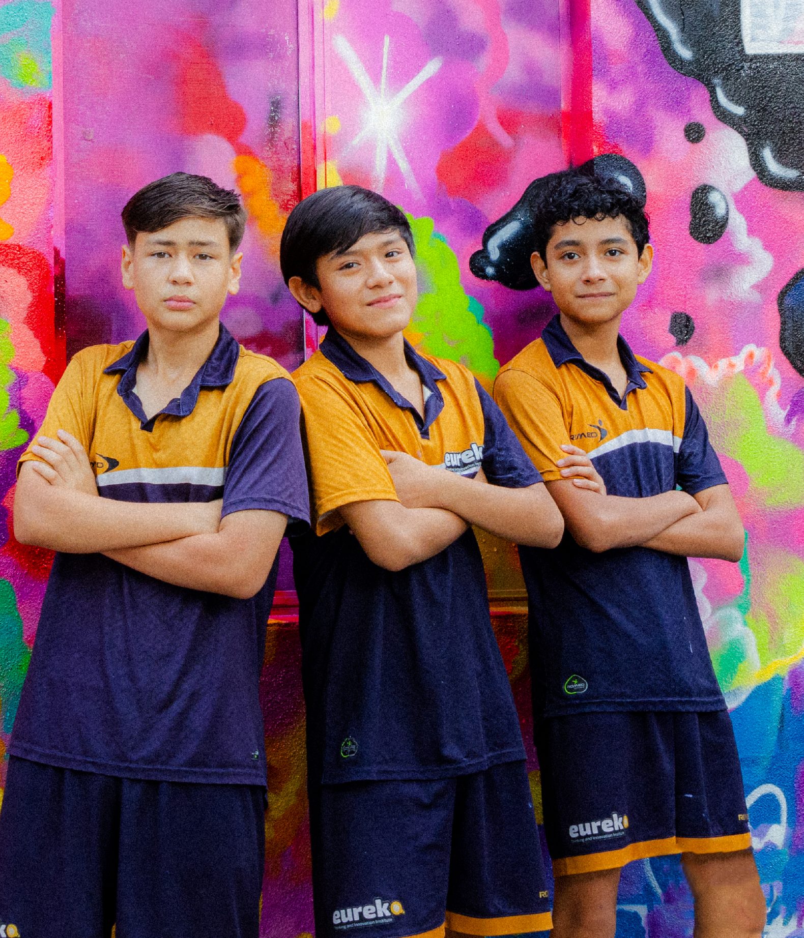 Tres alumnos de Eureka con el uniforme del instituto, de brazos cruzados frente a un mural de colores