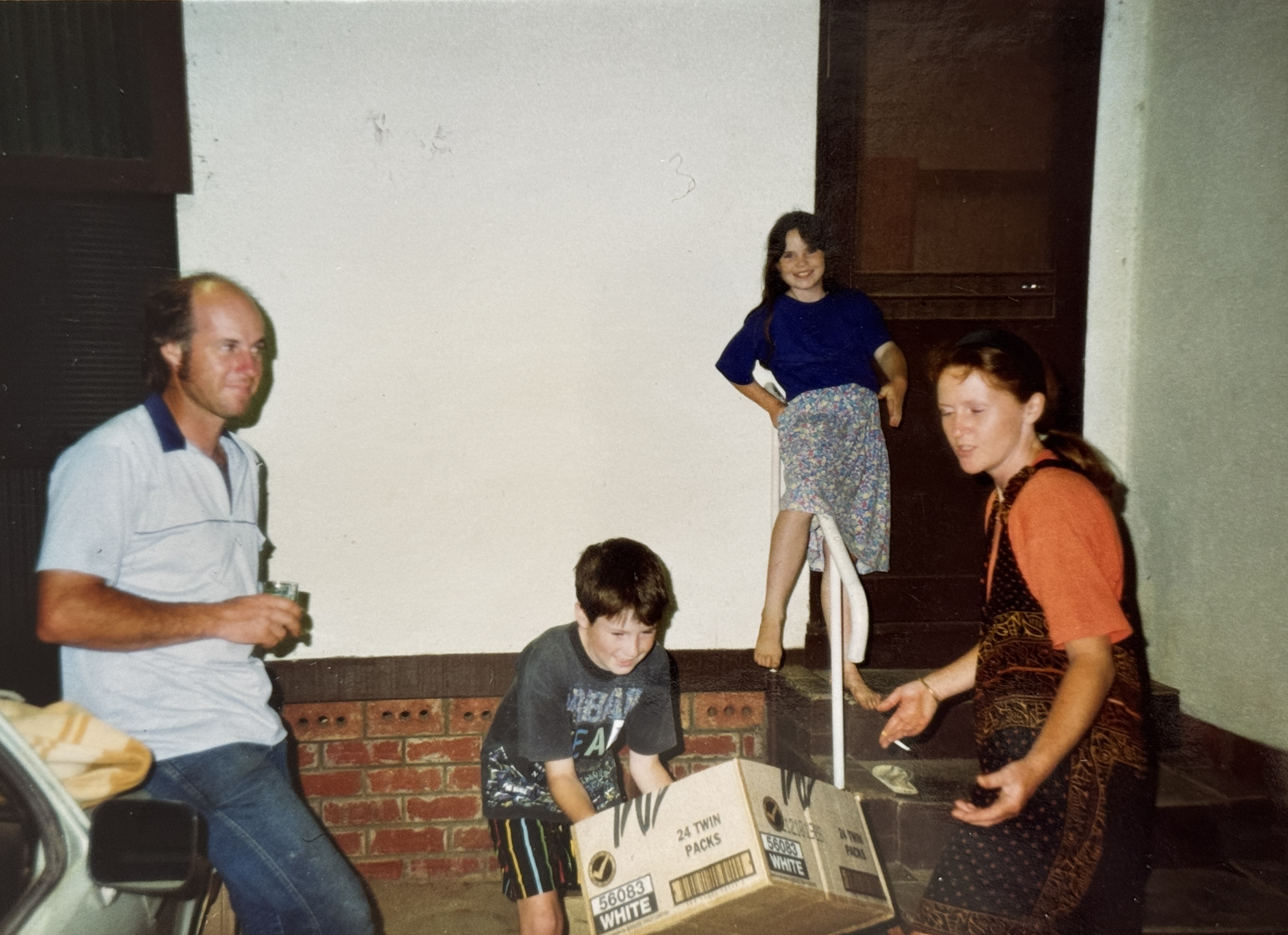 Family snapshot from the 1990s outside a brick home at night — a man leaning on a car, a boy playing with a cardboard box, a girl balancing barefoot on a step rail, and a woman laughing nearby — an ordinary moment that became a keepsake, echoing why you never get to choose when the photos stop. Family snapshot from the 1990s outside a brick home at night — a man leaning on a car, a boy playing with a cardboard box, a girl balancing barefoot on a step rail, and a woman laughing nearby — an ordinary moment that became a keepsake, echoing why you never get to choose when the photos stop.