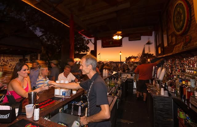 Busy outdoor bar at sunset, featuring patrons enjoying mojitos and cocktails, with a vibrant atmosphere reflecting Cuban culture.