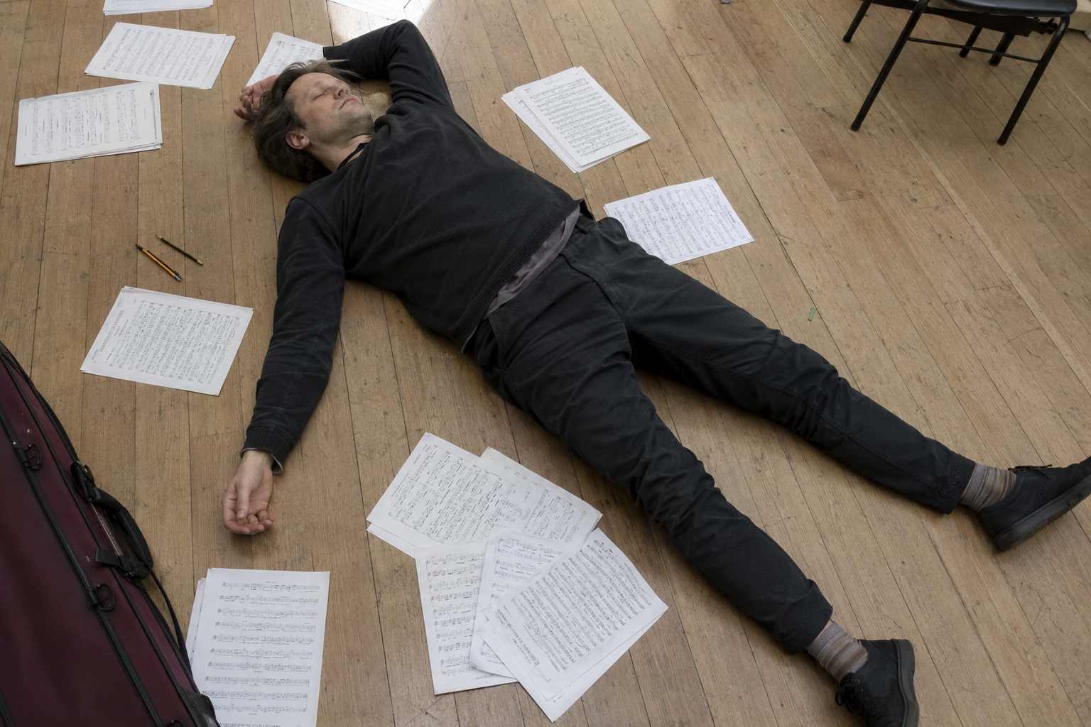 Exhausted musician lying on a wooden floor surrounded by scattered sheet music after an intense rehearsal session.