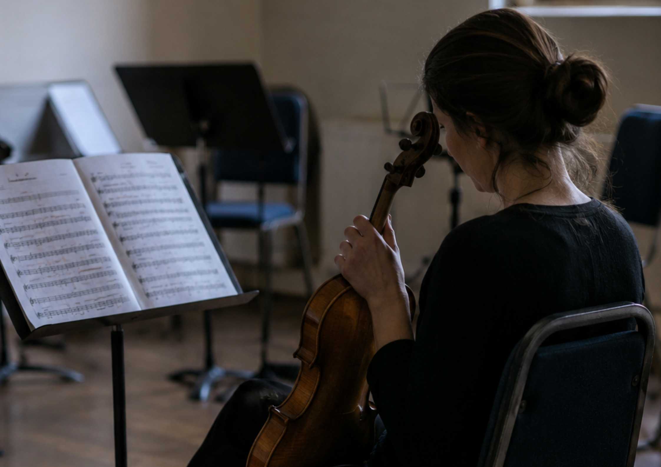 Female violinist sitting alone in an empty rehearsal room holding her violin, instrument case at her feet, music stand with score in front of her.