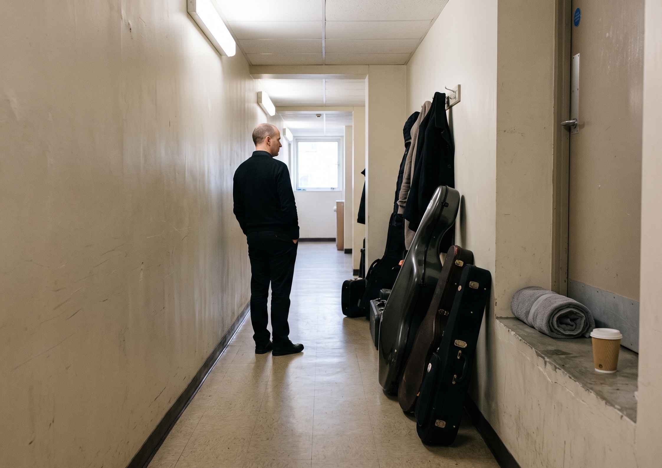 A backstage corridor with instrument cases along the wall and a musician standing further down with their back partly turned.