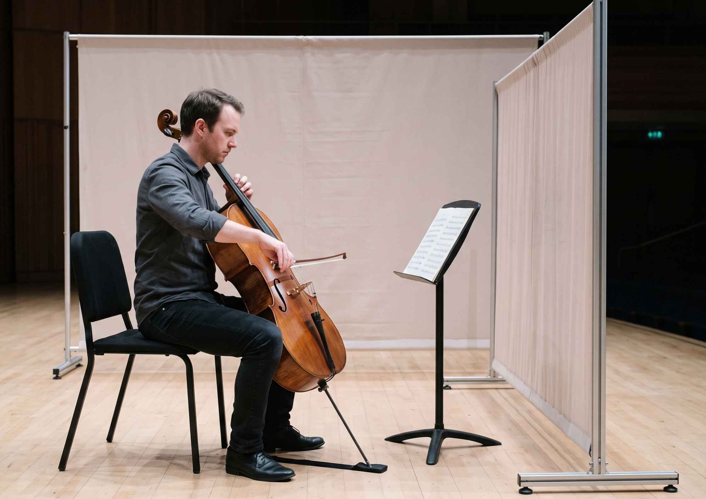 A cellist plays on a bare stage while a long fabric screen separates him from the dark, empty hall.