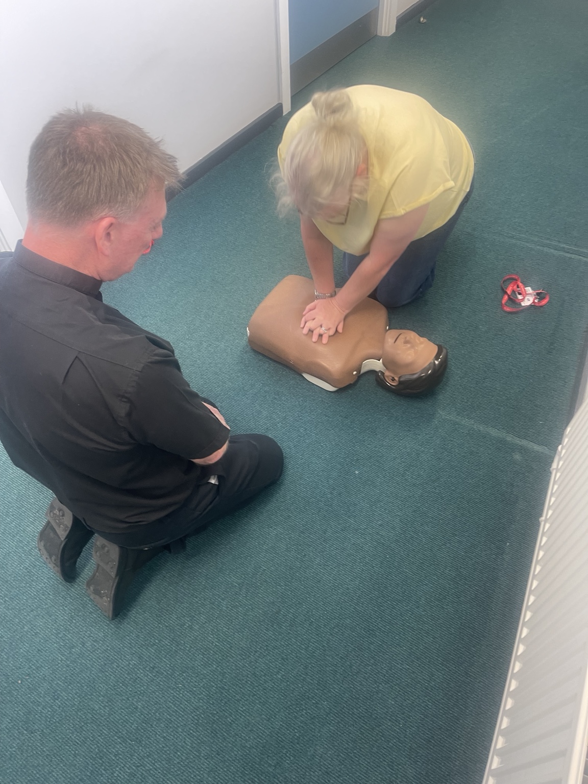 A trainer coaching a learner through adult CPR practice on a training manikin on the floor during a First Aid at Work course
