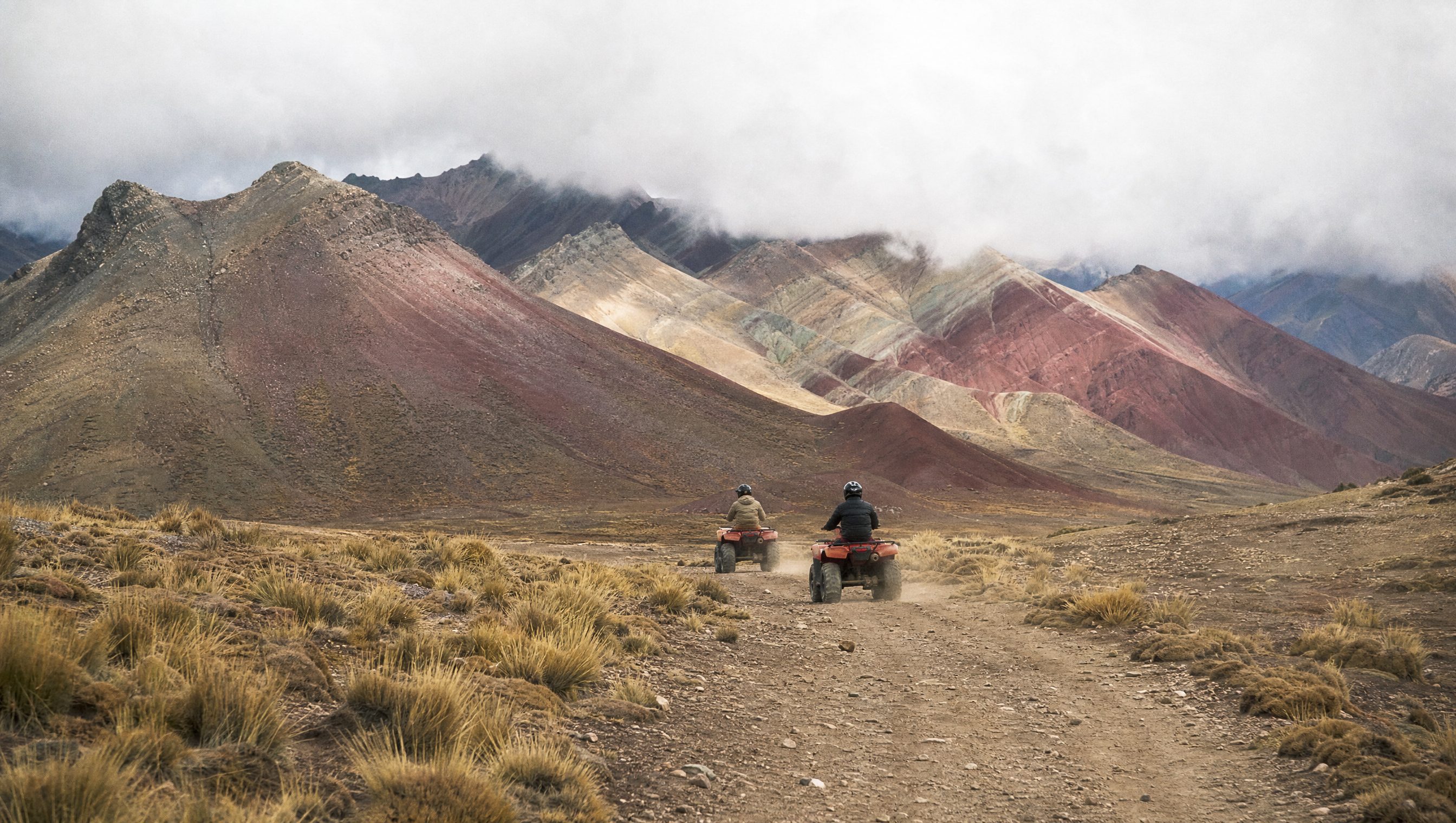 Two riders on quad bikes on a dirt track toward striped Andean mountains