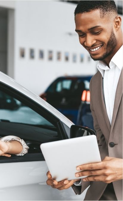 Smiling man in a suit showing a tablet to a customer in a car dealership. Highlights the effectiveness of Google ads and Facebook and Instagram ads in lead generation and getting more clients for local businesses in the automotive industry