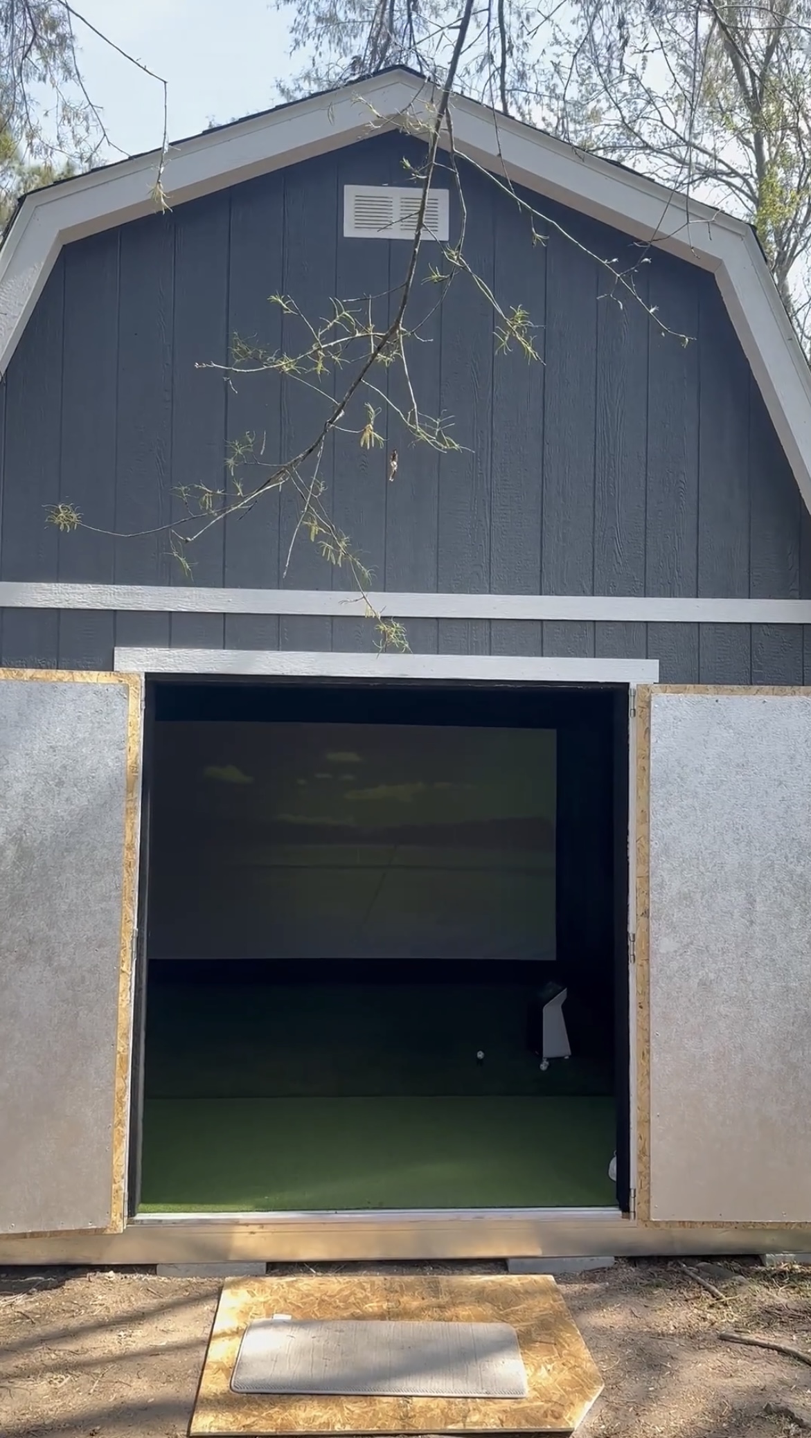 A real custom golf simulator shed in a wooded backyard, doors open to the turf and impact screen.