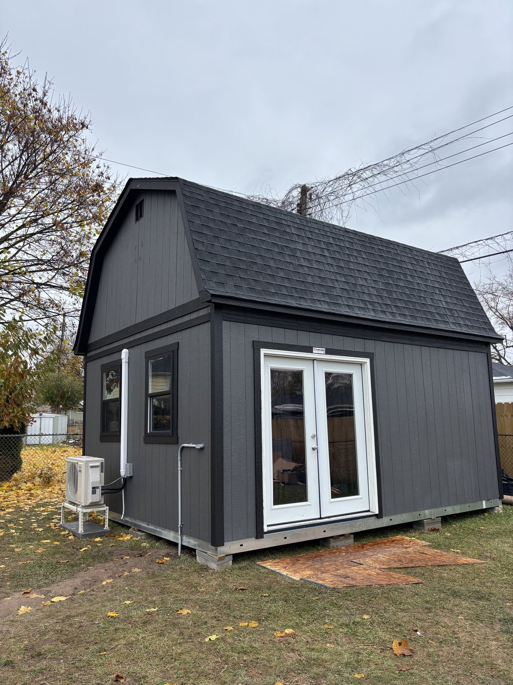The outside of a real customer golf simulator shed, dark gray with a gambrel roof and French doors, set in a backyard.