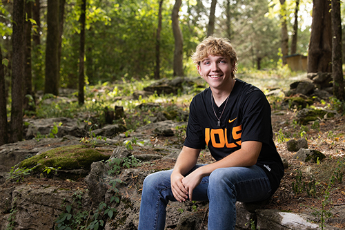 High school senior in a black T-shirt sitting on a stone wall among trees