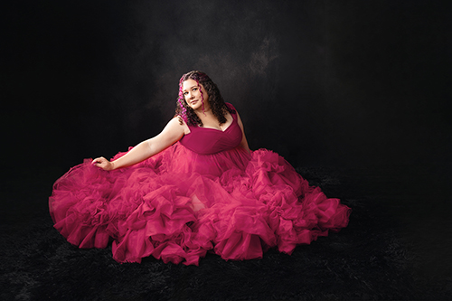A woman seated in a full burgundy gown against a dark studio backdrop.