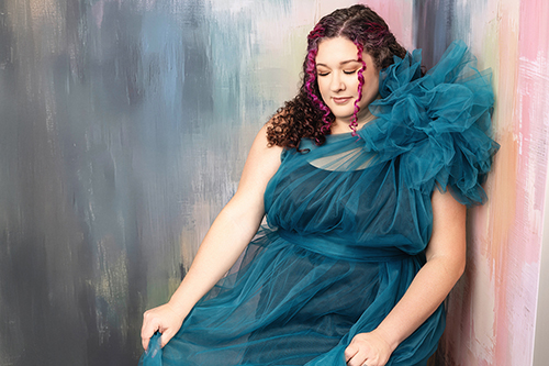 A woman in a teal dress looking down with a soft smile against a pale studio backdrop.
