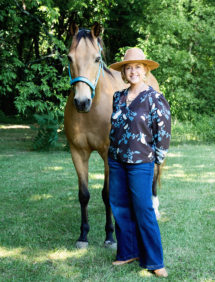 Angie Joseph smiling beside a horse outdoors, wearing a hat and floral blouse.