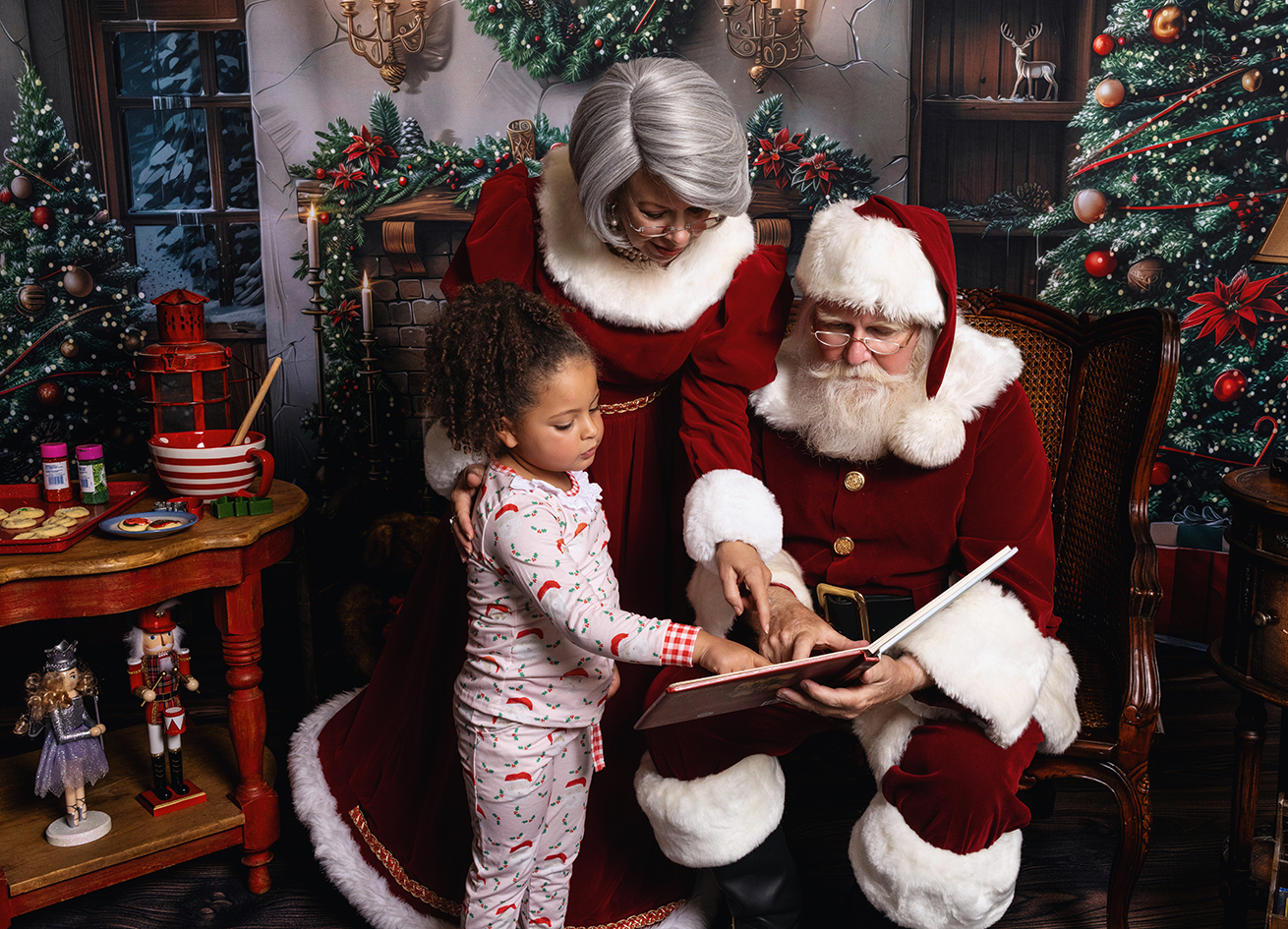 Young girl looking at an open book with Santa while Mrs. Claus leans beside them.