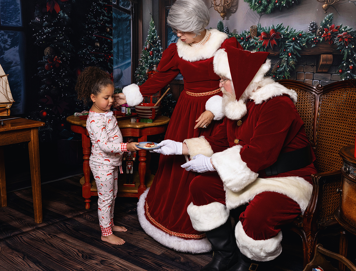 Young girl standing beside Mrs. Claus as Santa holds out a plate in the Christmas studio.