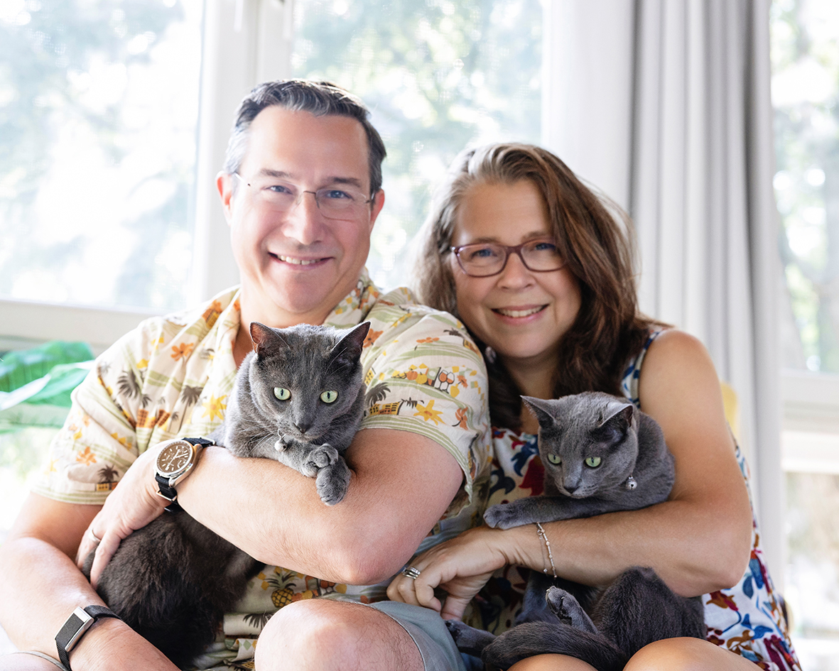 A smiling couple holding two gray cats beside a window