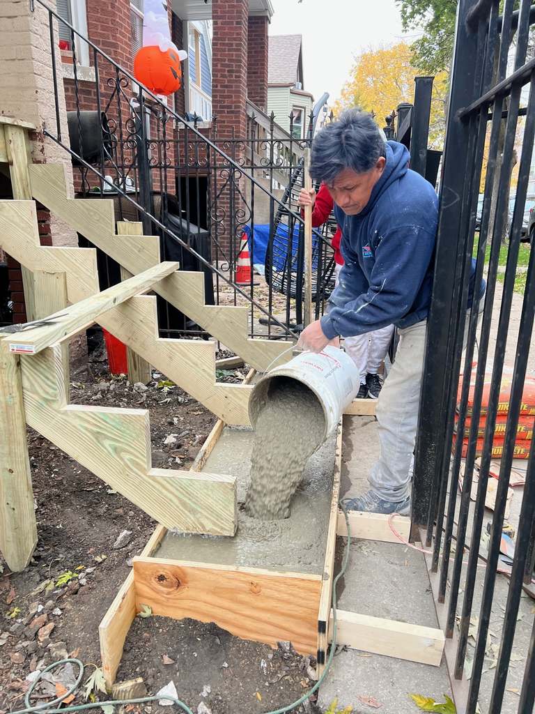 Wooden outdoor stair framing while a worker pours concrete from a bucket