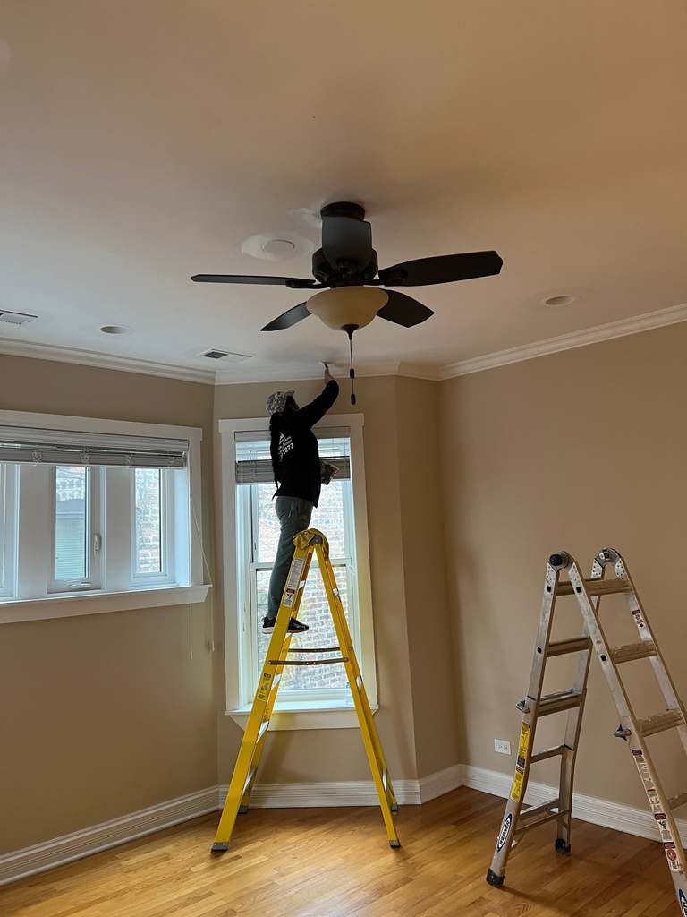 Worker on a yellow ladder next to a ceiling fan in a room with large windows