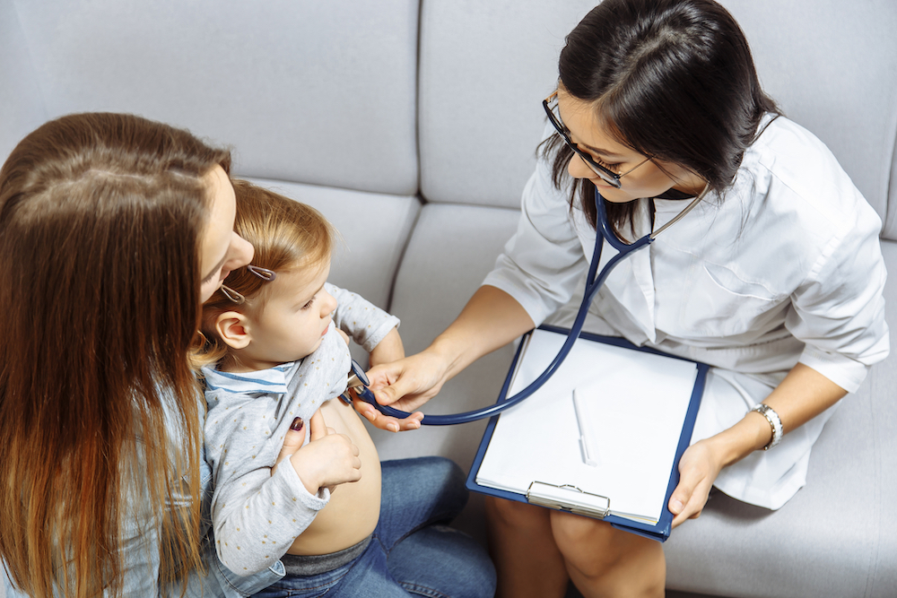 A clinician listening to a young child&rsquo;s chest with a stethoscope while a parent holds them