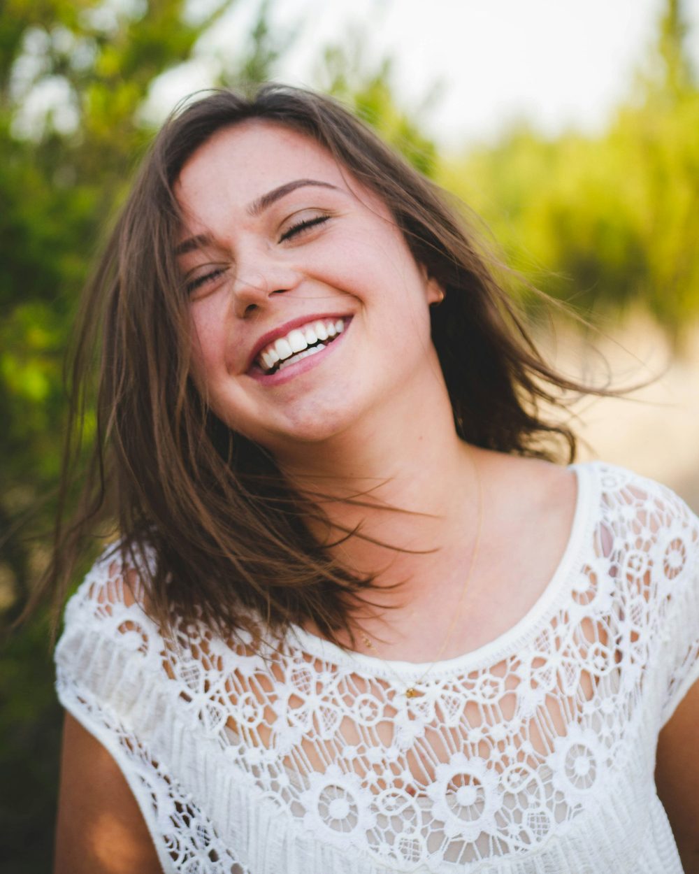A woman laughing outdoors with green trees behind her