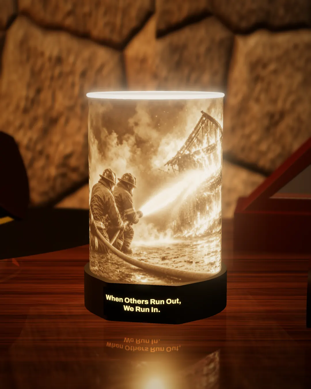 Firefighter tribute lithophane lamp lit warm, showing firefighters working a hose line into the flames, with the black base nameplate glowing “When Others Run Out, We Run In.”, on a wooden desk against a stone wall