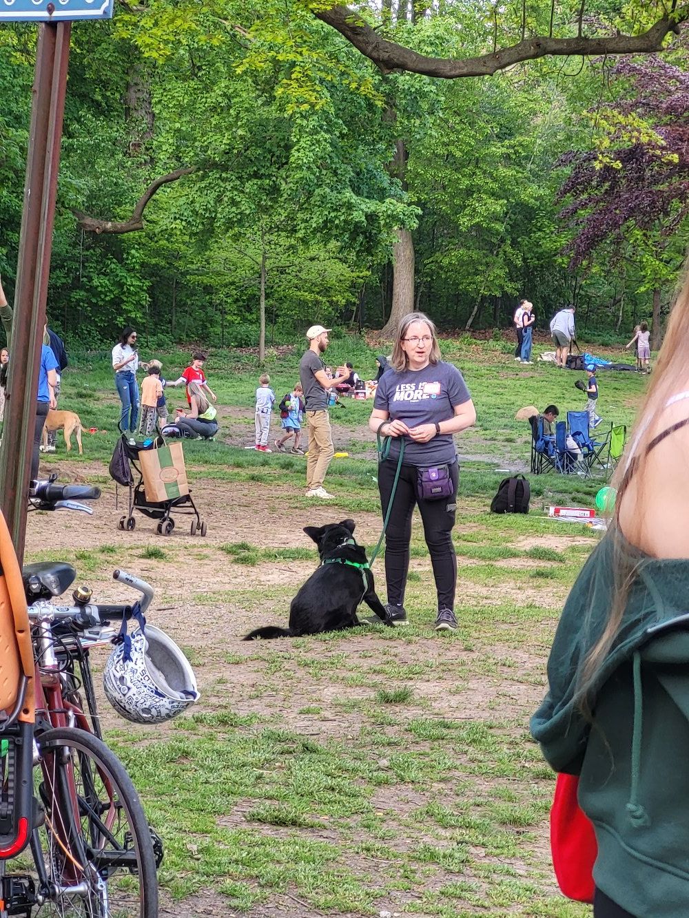 A woman (me) in a t-shirt & athletic pants with a treat pouch on stands in a crowded park. She holds a leash. A medium-sized black dog in a harness is on the leash and sitting at her feet, scratching his ear with his back foot.
