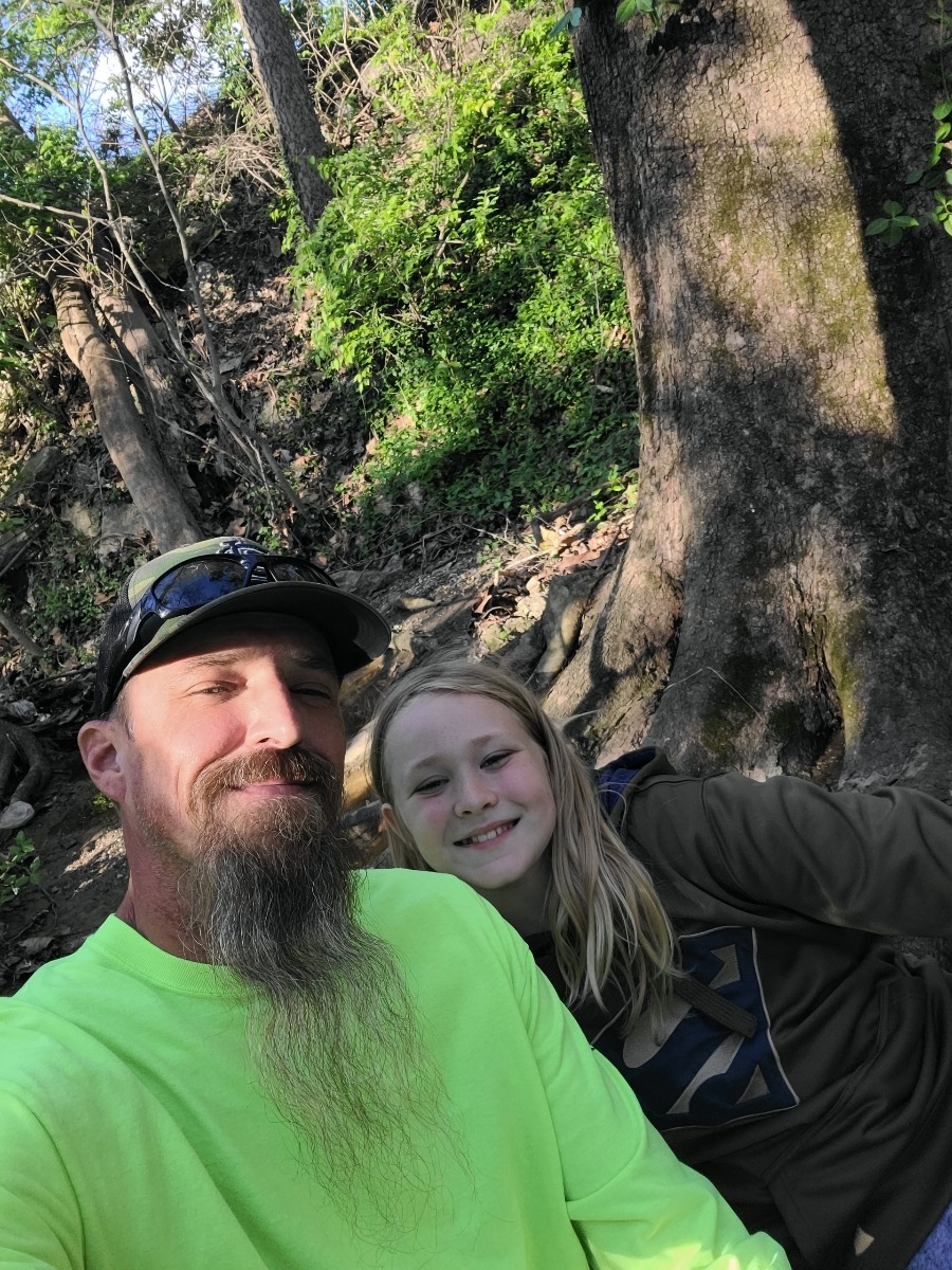 Man and young girl smiling while resting on a wooded hiking trail beside large trees.