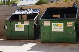 Two large green industrial dumpsters from Waste Management with open lids stand side-by-side on concrete.