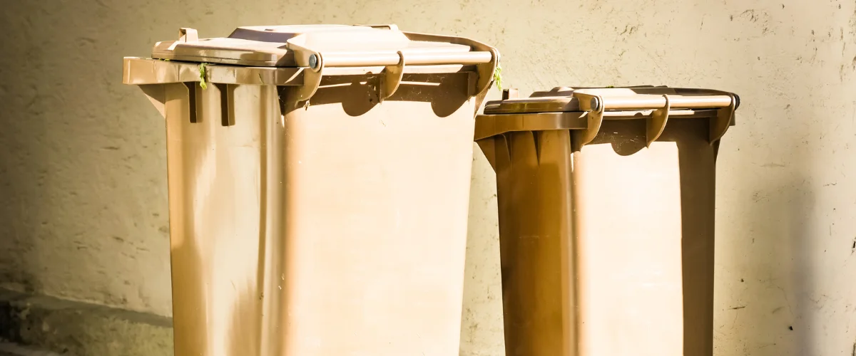 Two brown plastic recycling bins, one larger than the other, standing against a beige wall in sunlight. Two brown plastic recycling bins, one larger than the other, standing against a beige wall in sunlight.