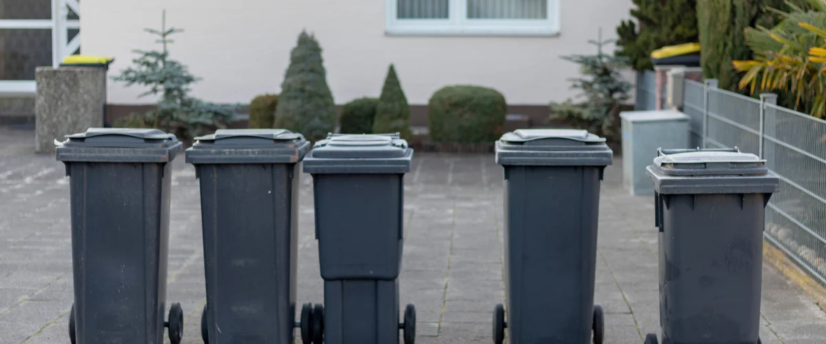 A row of five dark gray trash cans with lids, lined up on a paved surface in front of a house. A row of five dark gray trash cans with lids, lined up on a paved surface in front of a house.