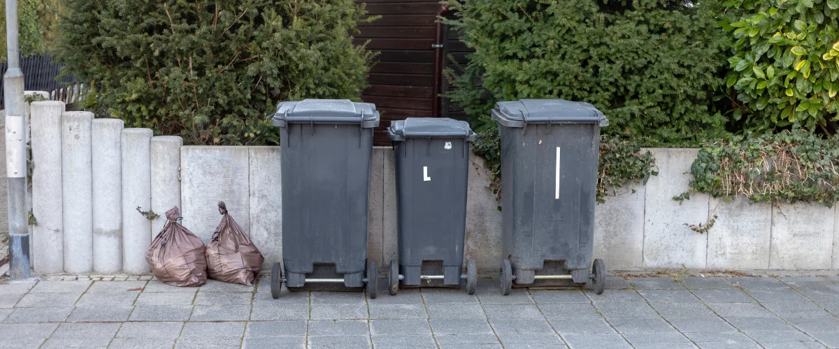 Three gray plastic wheelie bins of different sizes, including one marked 'L', stand on a paved street beside a concrete wall and two brown garbage bags. Three gray plastic wheelie bins of different sizes, including one marked 'L', stand on a paved street beside a concrete wall and two brown garbage bags.