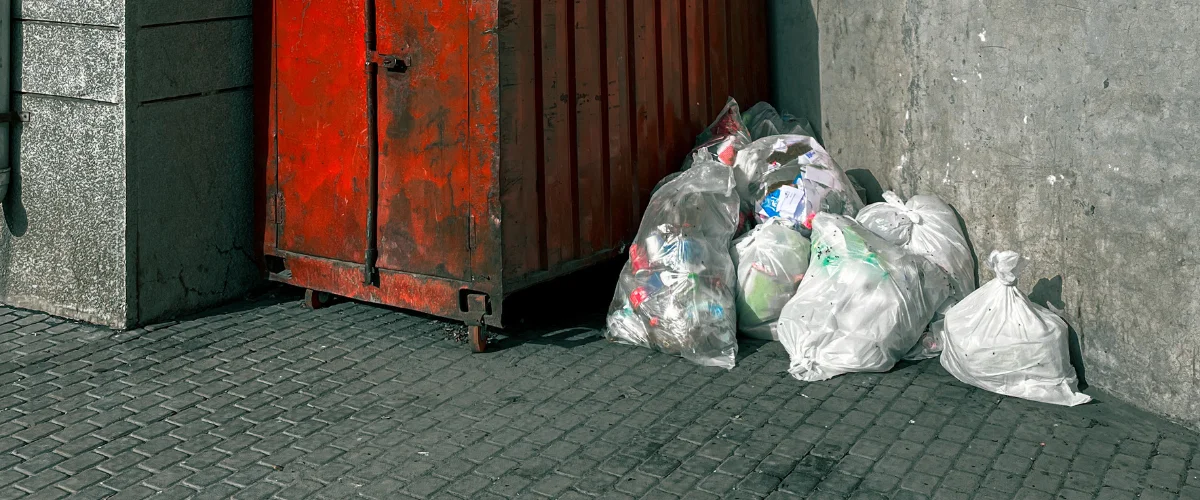 A weathered red dumpster next to a concrete wall with several transparent and white trash bags filled with refuse. A weathered red dumpster next to a concrete wall with several transparent and white trash bags filled with refuse.