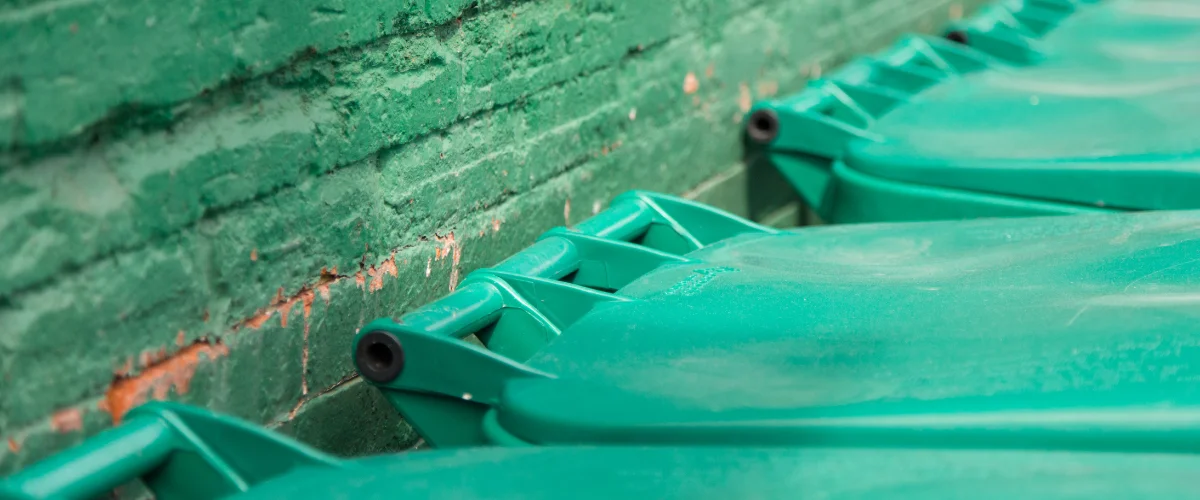 Close-up of rows of green plastic trash bins or containers against a textured green brick wall.