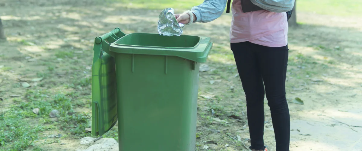 A person, partially visible, dropping crumpled aluminum foil into an open green public waste bin in a park setting. A person, partially visible, dropping crumpled aluminum foil into an open green public waste bin in a park setting.