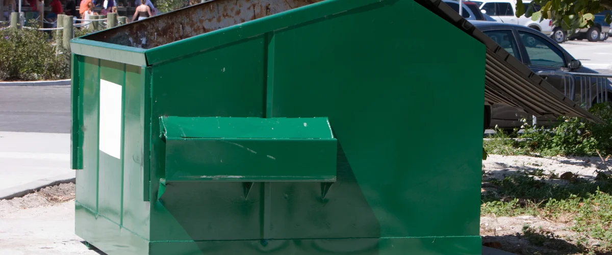 A large, weathered green industrial dumpster with a small side opening, sitting on a paved surface in daylight.
