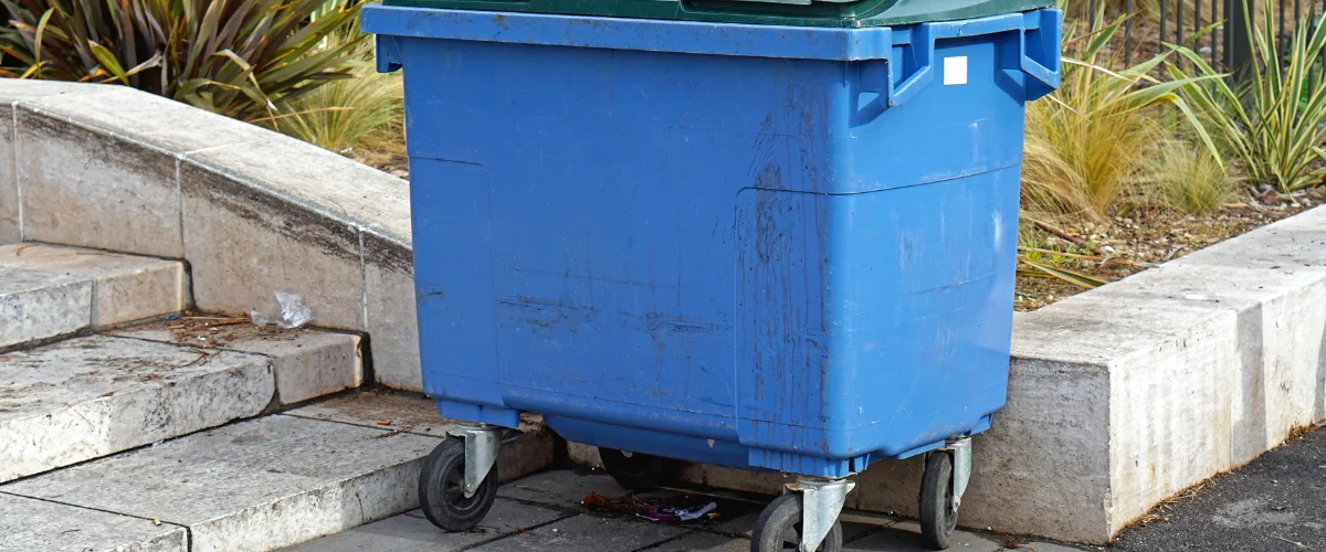 A large blue dumpster with a green lid on wheels, sitting on a paved surface next to concrete steps and a garden.