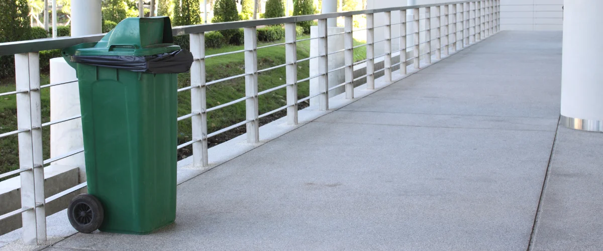 A green wheeled trash bin with a black liner on a concrete walkway next to a modern metal railing. A green wheeled trash bin with a black liner on a concrete walkway next to a modern metal railing.