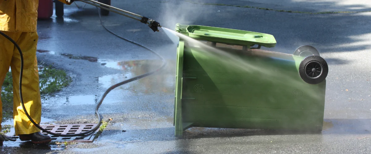 A person in yellow rain gear pressure washes a green wheelie bin lying on its side on wet asphalt. A person in yellow rain gear pressure washes a green wheelie bin lying on its side on wet asphalt.