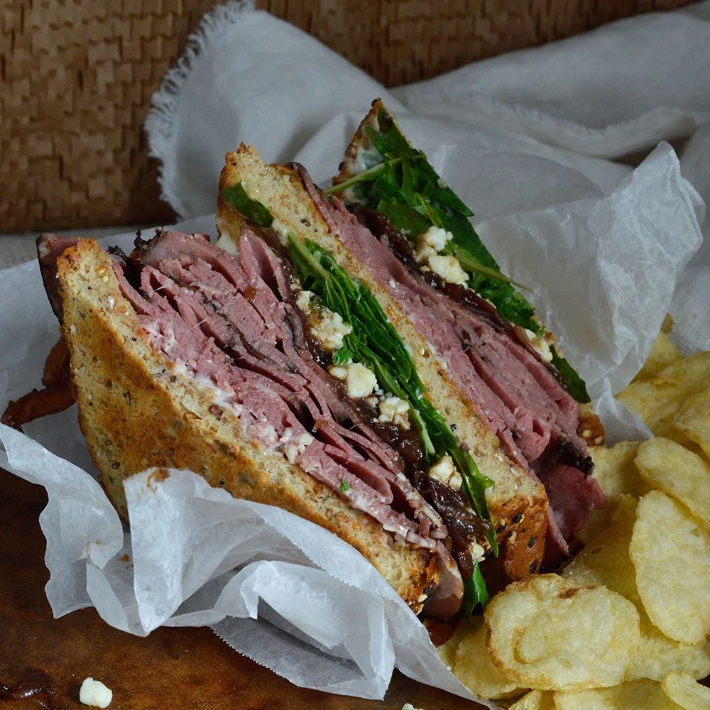 A deli sandwich stacked with sliced roast beef, greens and crumbled cheese, served with chips
