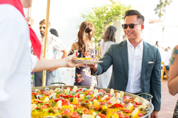 Server handing a plate of paella to a guest beside a large paella pan at a corporate event