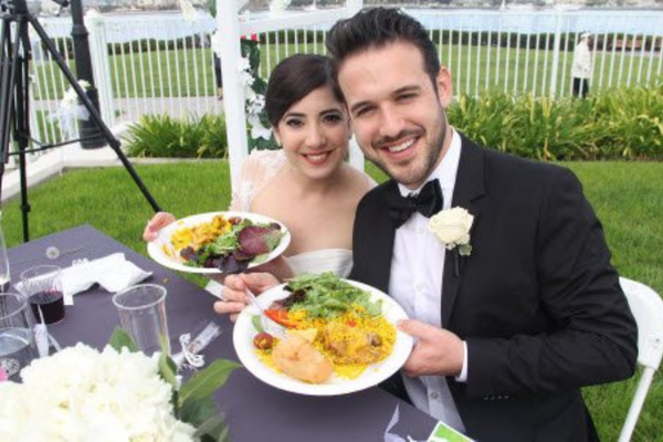 Bride and groom holding plates of paella at an outdoor wedding reception