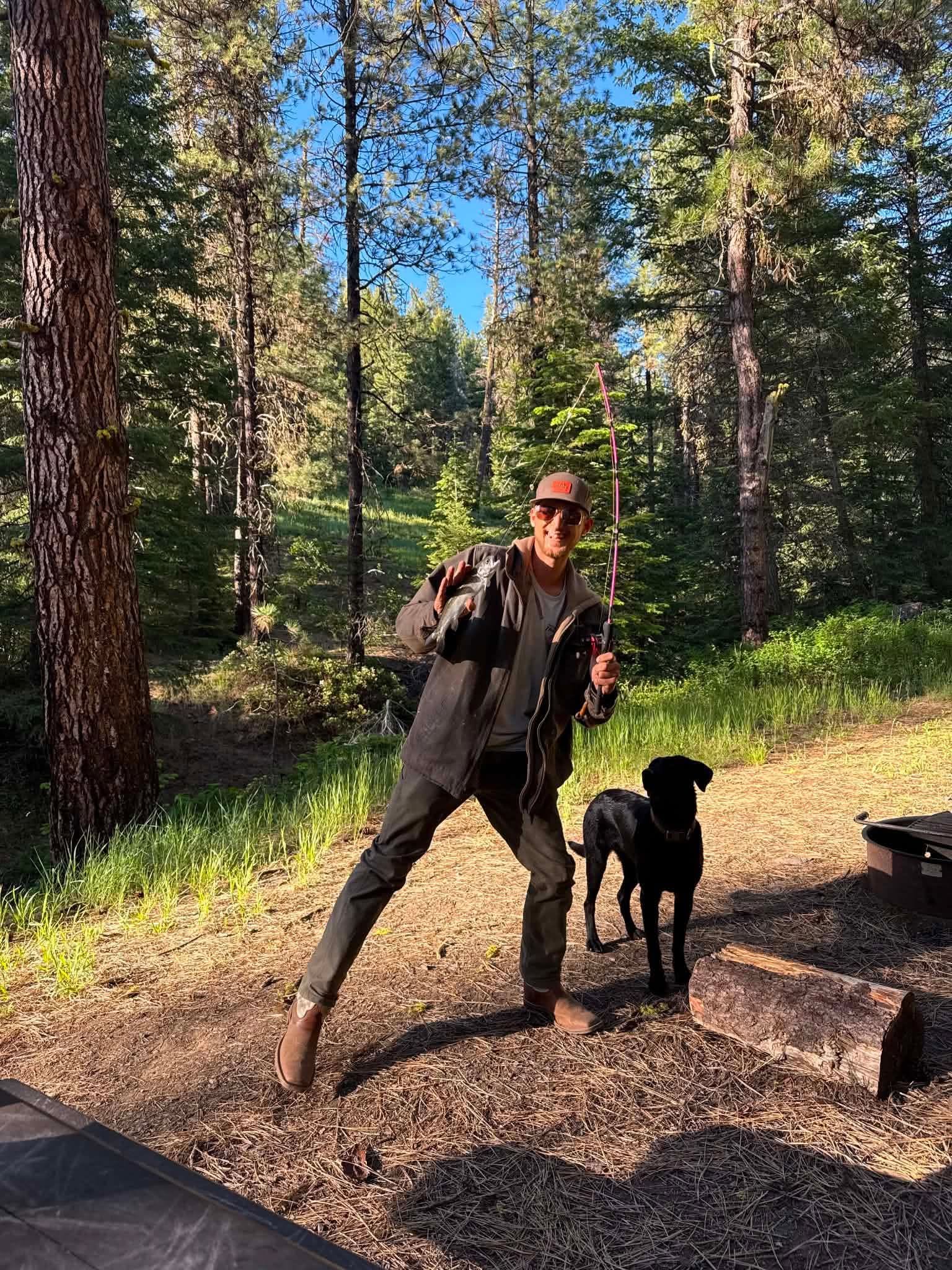 Daniel holding a fish and a pink Compensator rod beside his dog in the forest