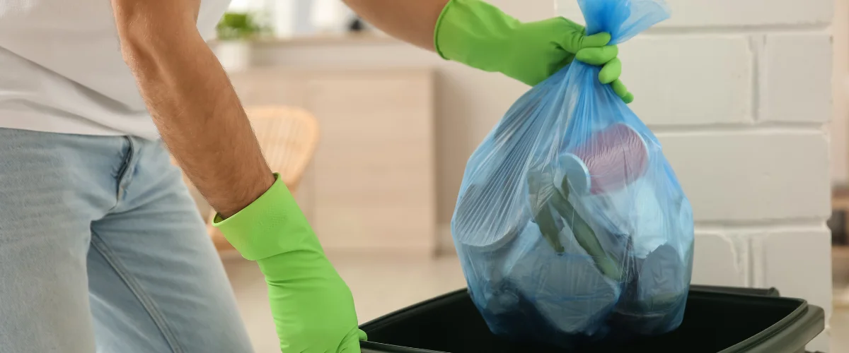 A person wearing green gloves holds a blue trash bag full of waste over a dark green bin, ready to dispose of it. A person wearing green gloves holds a blue trash bag full of waste over a dark green bin, ready to dispose of it.