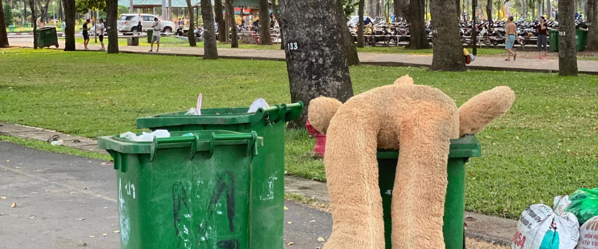 A large, light brown teddy bear draped upside down over a green trash can in a park setting.