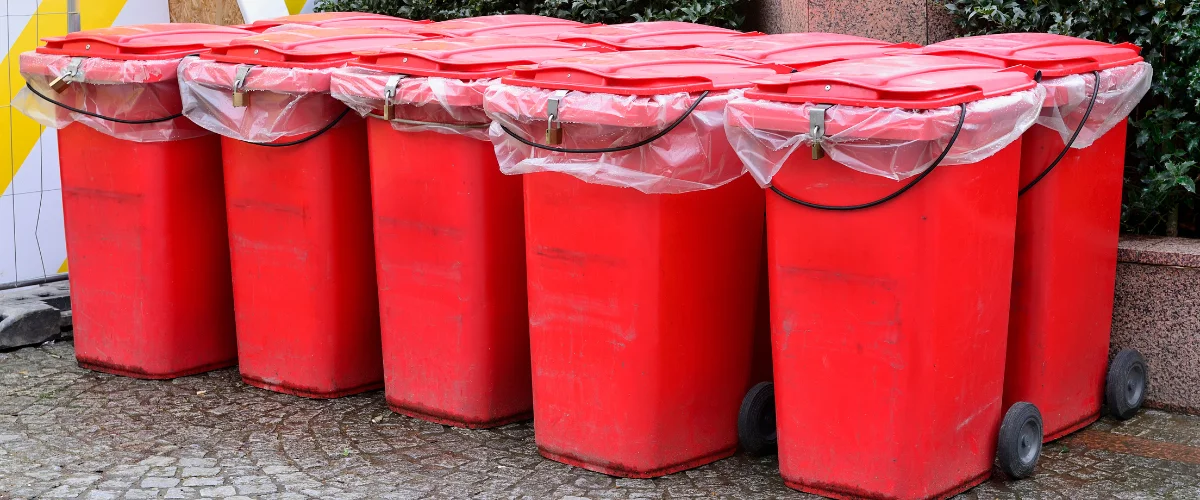 A line of six bright red garbage bins with plastic bags and locks on their lids, sitting on a cobblestone street.