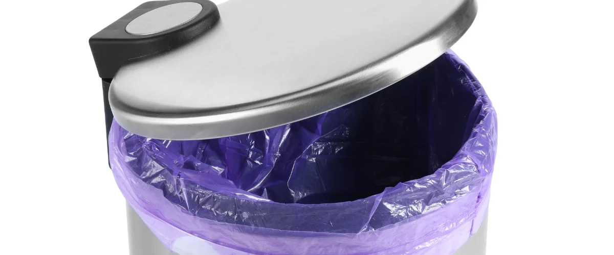 A close-up shot of a stainless steel trash can with an open lid, revealing a vibrant purple bin liner inside.